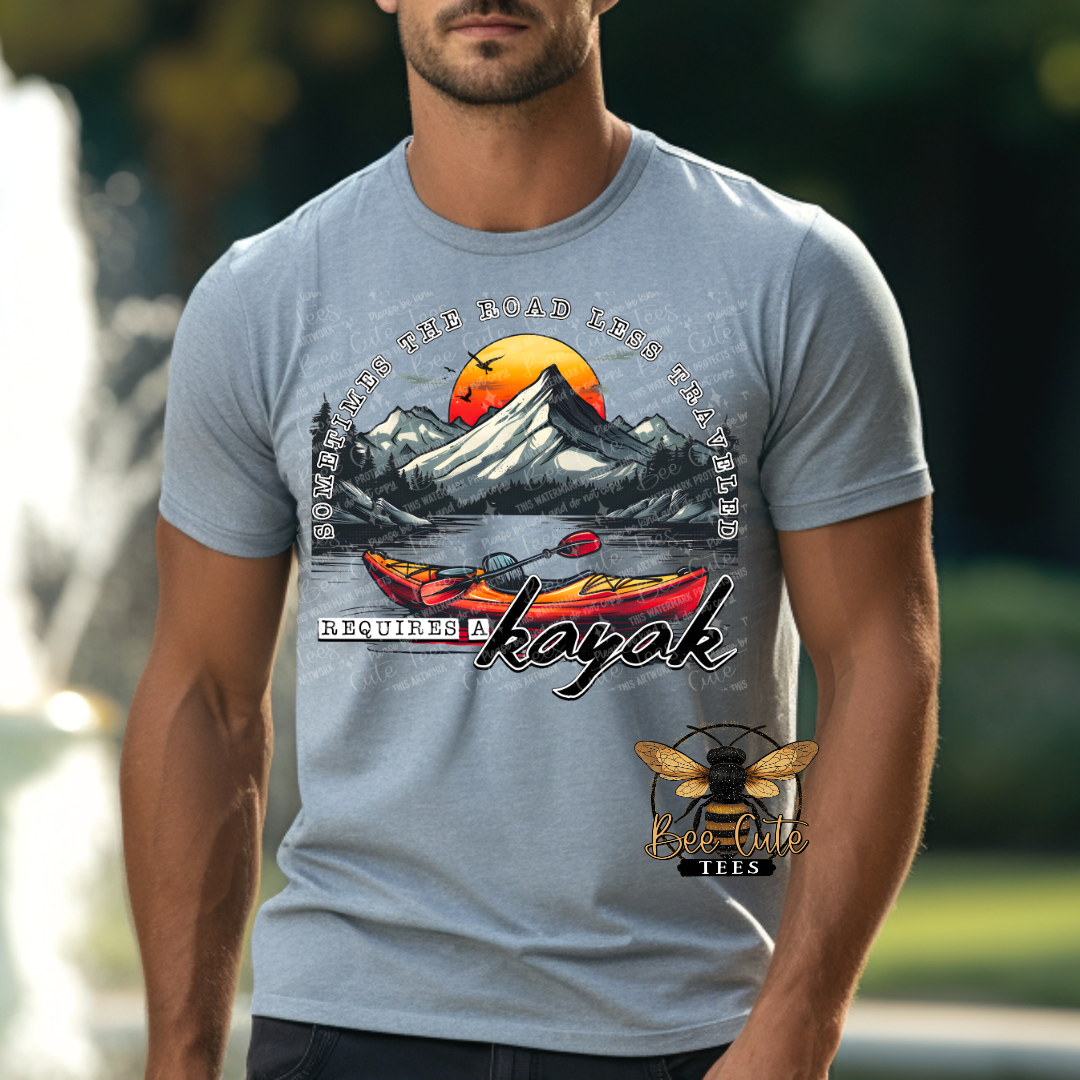 Man wearing a grey graphic tee featuring colorful kayaks on a lake with mountain scenery and a sunset background, paired with the phrase “Sometimes the Road Less Traveled Requires a Kayak.”
