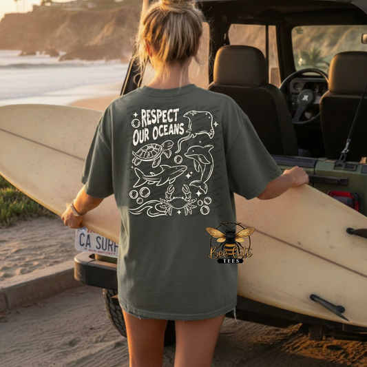 Person holding a surfboard wearing a t-shirt with ocean-themed design and text on a beach.