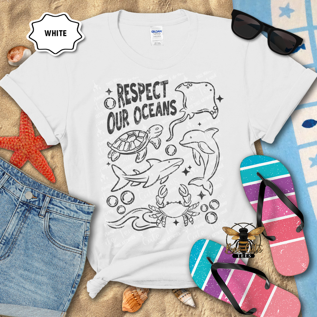 White t-shirt with ocean-themed design and beach accessories on a sandy background
