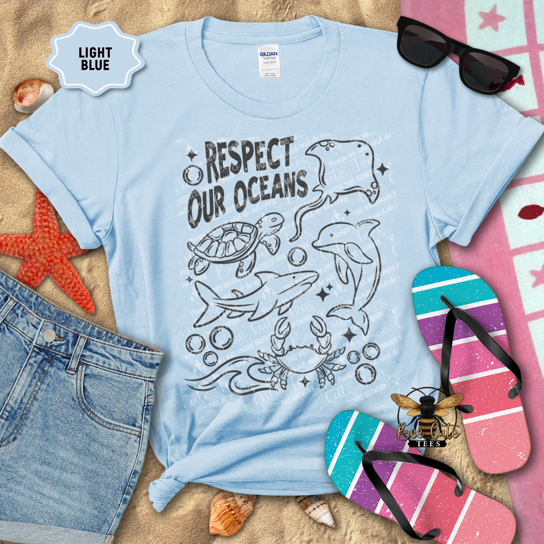 Light blue t-shirt with ocean-themed design and beach accessories on a sandy background