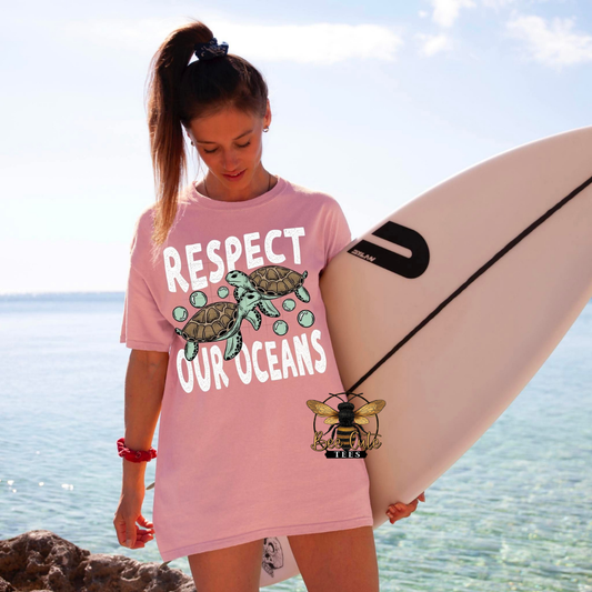 Woman holding a surfboard wearing a pink 'Respect Our Oceans' t-shirt with turtle graphics on a beach.