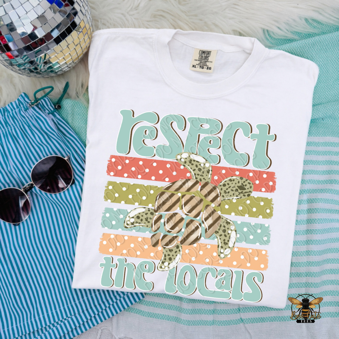 Folded t-shirt with 'Respect the Locals' design on a textured surface with sunglasses and a disco ball.