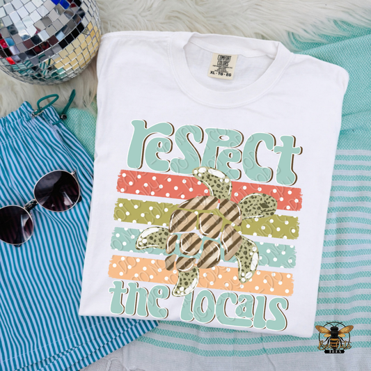 Folded t-shirt with 'Respect the Locals' design on a textured surface with sunglasses and a disco ball.