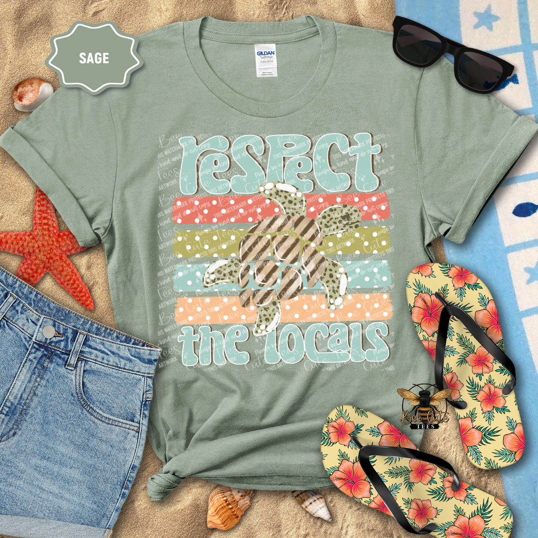 Sage green t-shirt with 'Respect the Locals' graphic on a beach setting with flip-flops and sunglasses.