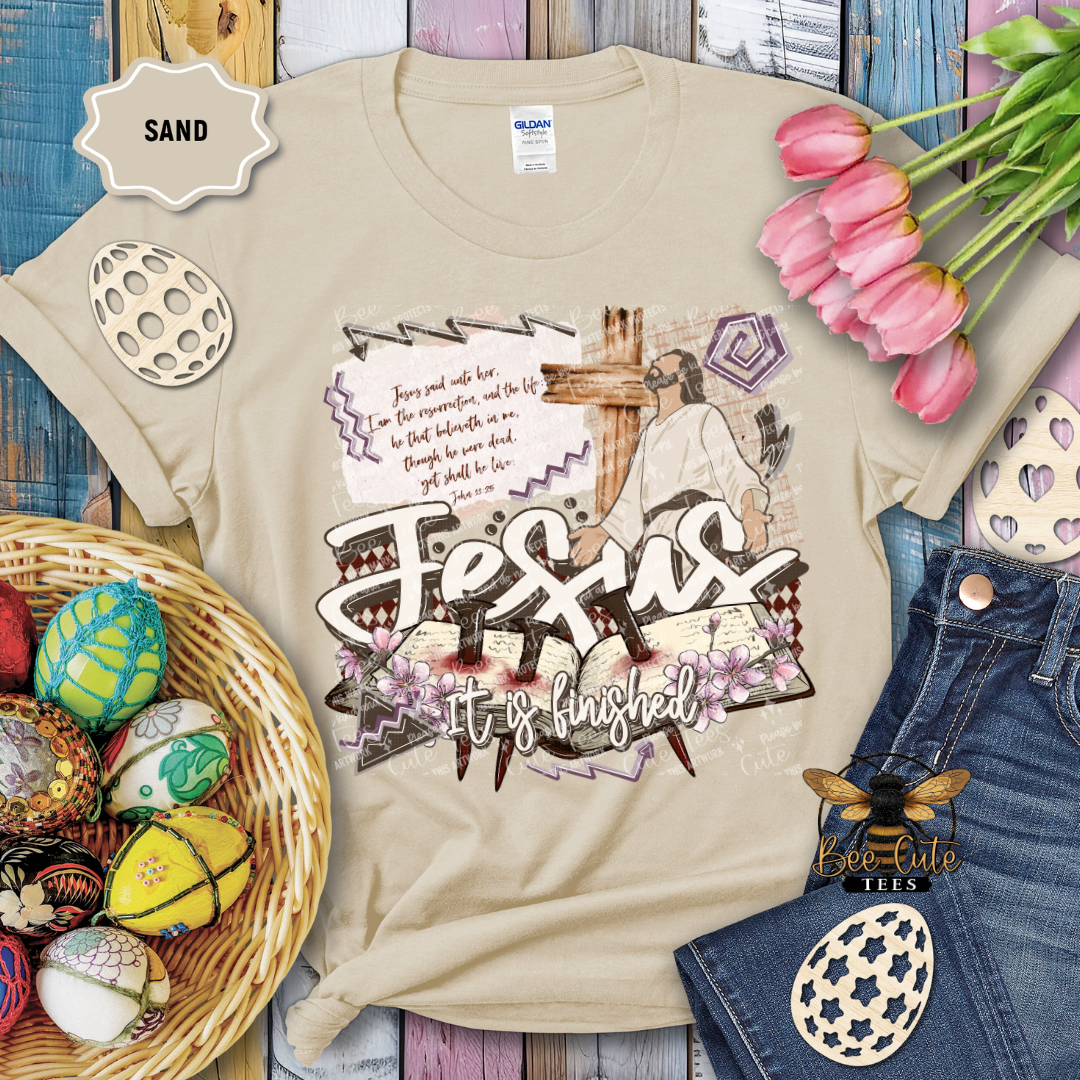 Beige t-shirt with 'Jesus It's Finished' design, surrounded by Easter decorations on a wooden surface.
