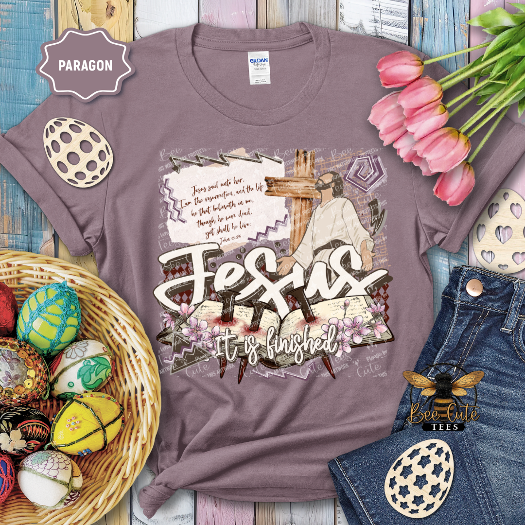 Purple t-shirt with 'Jesus it's finished' design, surrounded by Easter decorations on a wooden surface.