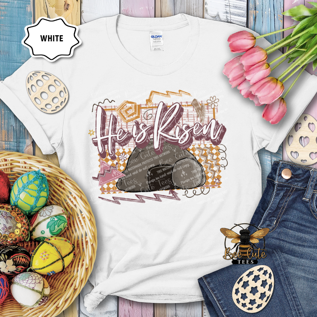 White t-shirt with 'He's Risen' design, surrounded by Easter decorations on a wooden surface.
