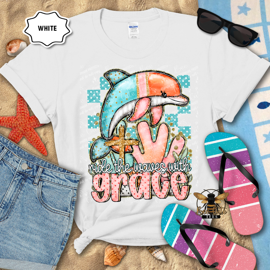 White t-shirt with a colorful dolphin design and 'ride the waves with grace' text, surrounded by beach accessories.