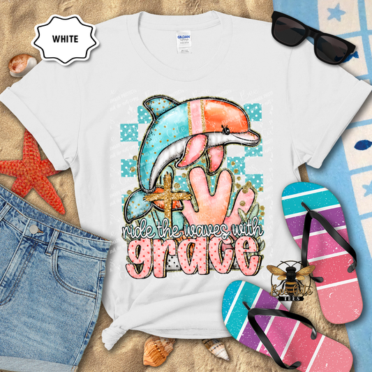 White t-shirt with a colorful dolphin design and 'ride the waves with grace' text, surrounded by beach accessories.