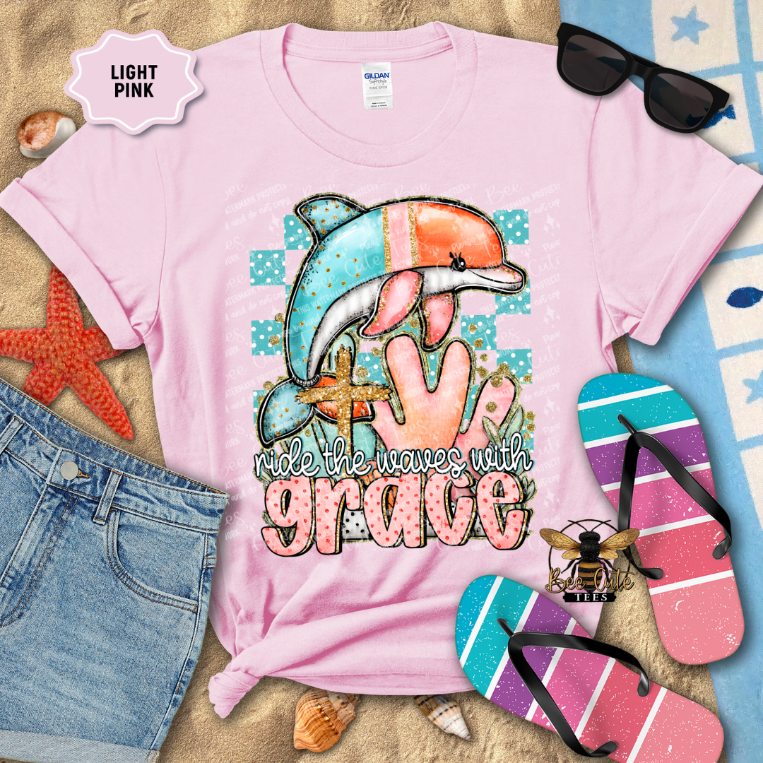 Light pink t-shirt with a dolphin graphic and 'ride the waves with grace' text, surrounded by beach accessories.