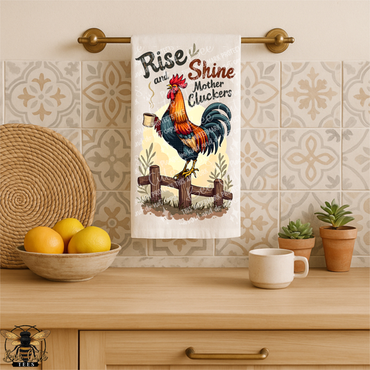 Kitchen towel with rooster design and text hanging on a towel rack, with a bowl of lemons, mug, and plants in the background.