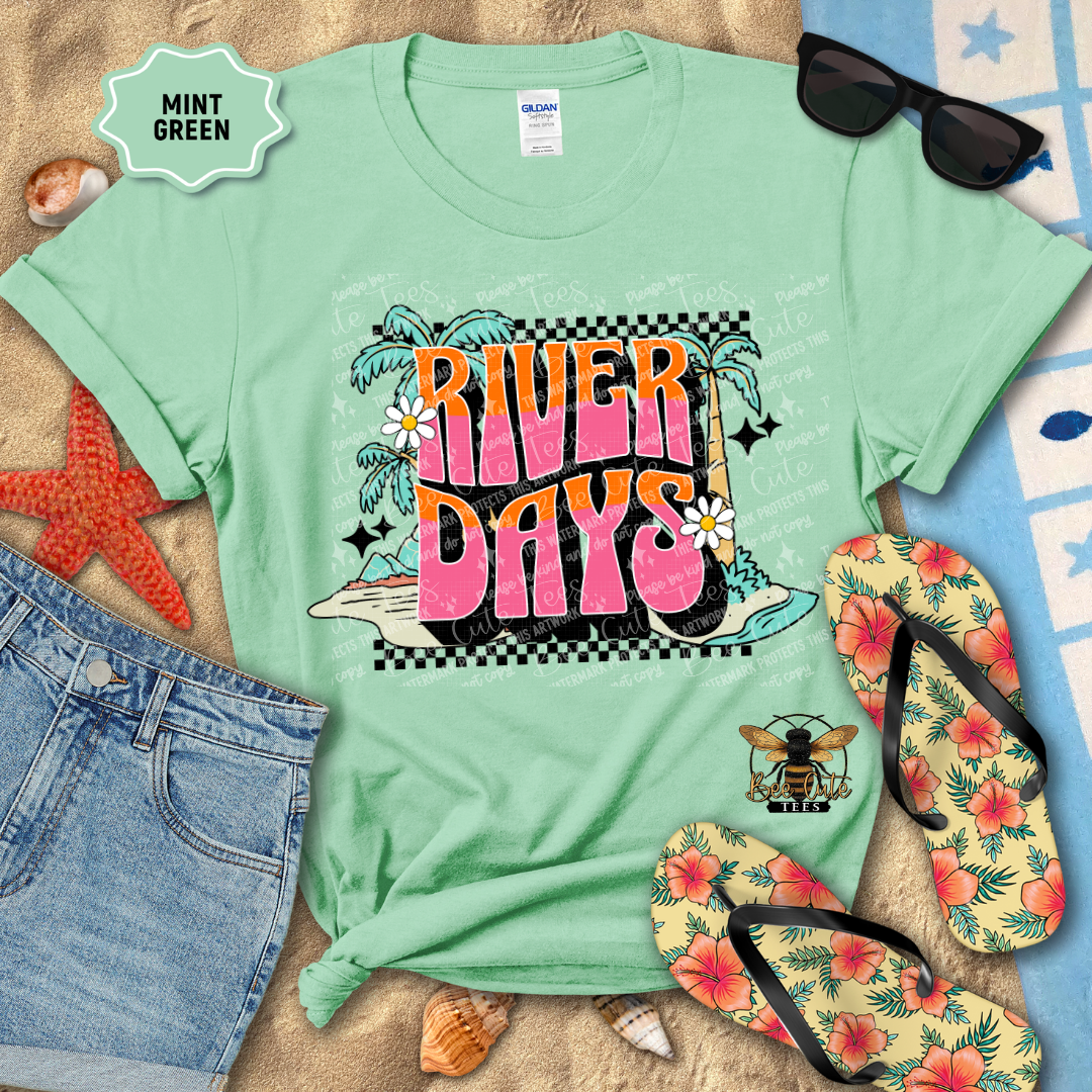 Mint green t-shirt with 'River Days' design on sand with beach accessories.