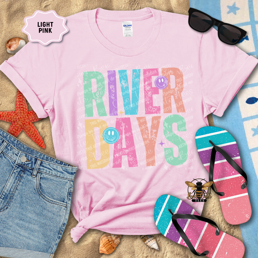 Pink t-shirt with 'River Days' text surrounded by beach accessories on a sandy background