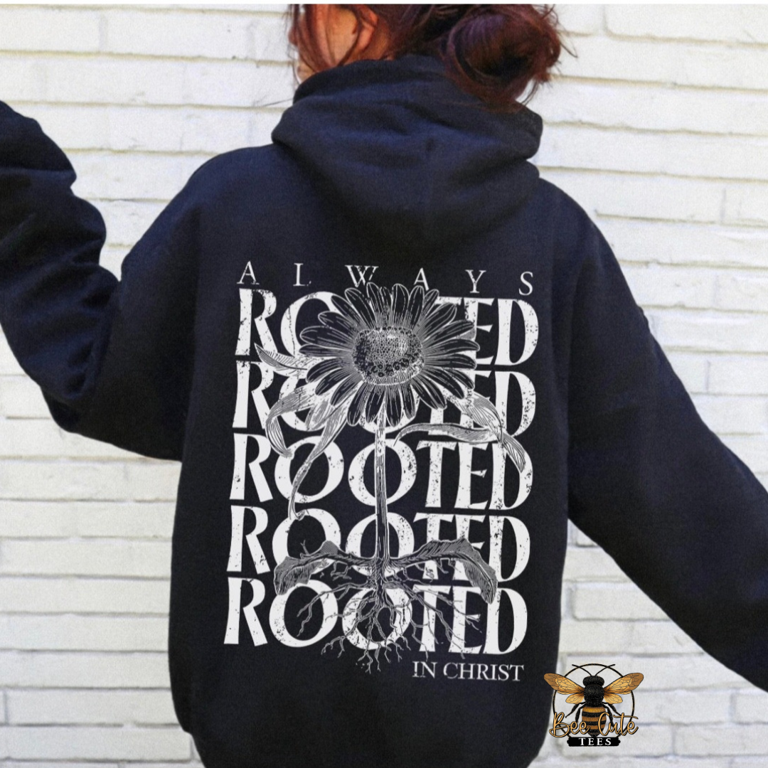 A person wearing a black hoodie from the back, featuring a large white graphic print with a sunflower and the words 'ALWAYS ROOTED IN CHRIST'. The person is standing against a white brick wall.