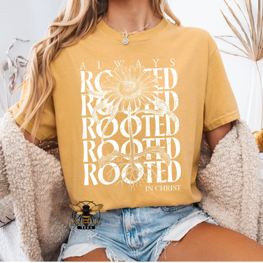 A woman wearing a mustard yellow graphic t-shirt with a white sunflower and the text 'ALWAYS ROOTED IN CHRIST'. She is also wearing denim shorts and a fuzzy beige cardigan.