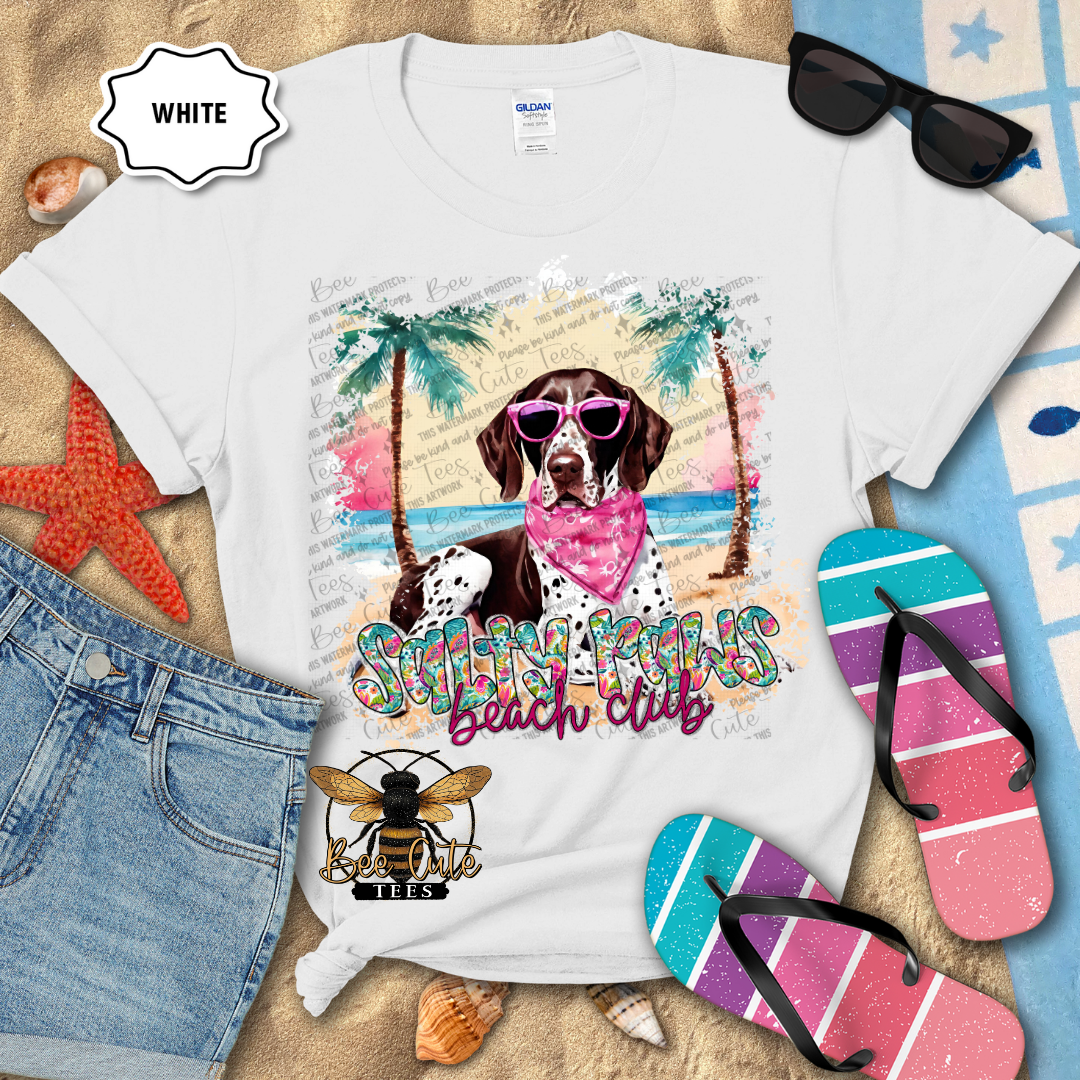 White t-shirt with a beach-themed design featuring dogs, sunglasses, and flip-flops on a sandy background.