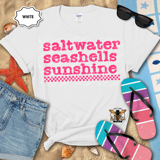 White t-shirt with 'saltwater seashells sunshine' text, sunglasses, flip-flops, and beach items on sand.