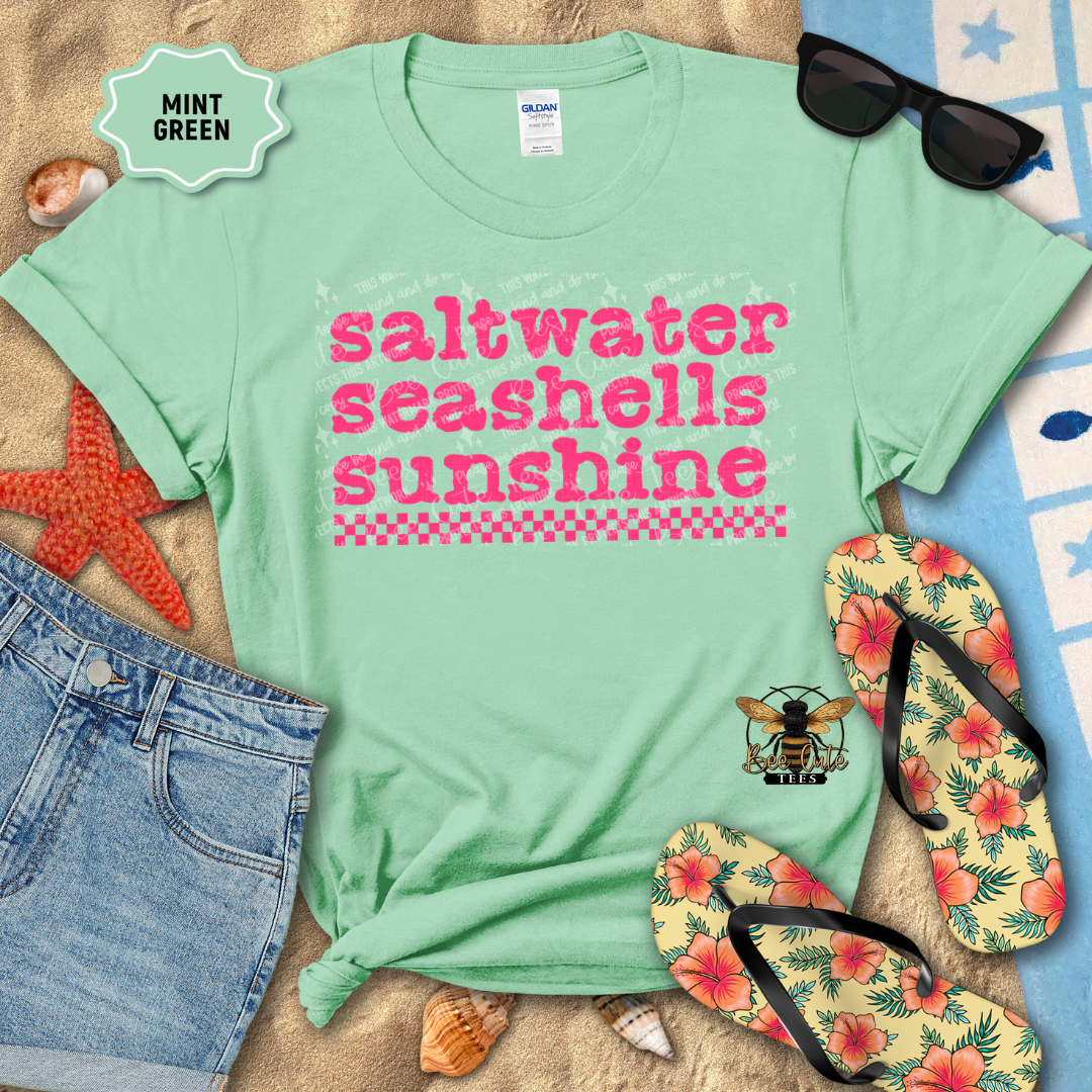 Mint green t-shirt with 'saltwater seashells sunshine' text, surrounded by beach accessories on sand.