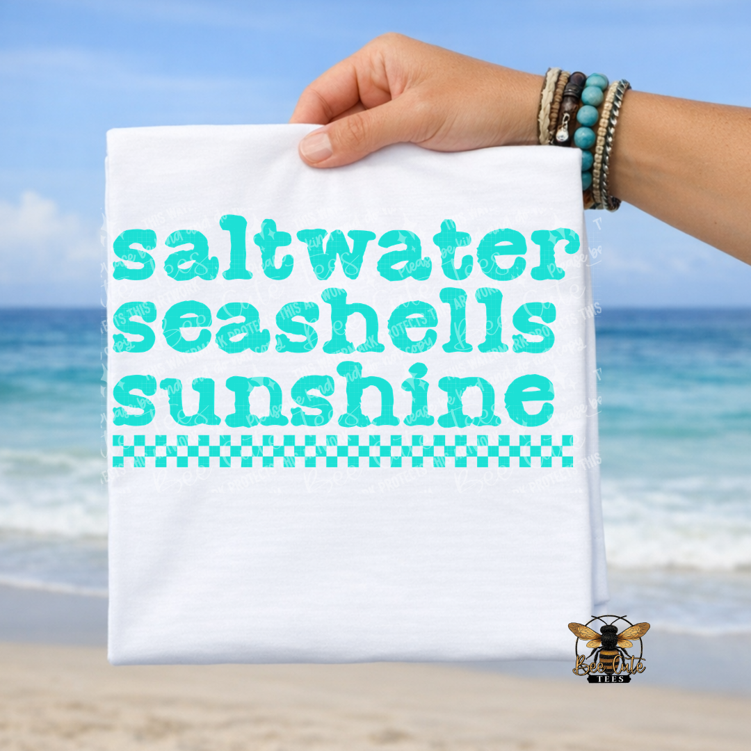Hand holding a t-shirt with 'saltwater seashells sunshine' text against a beach background