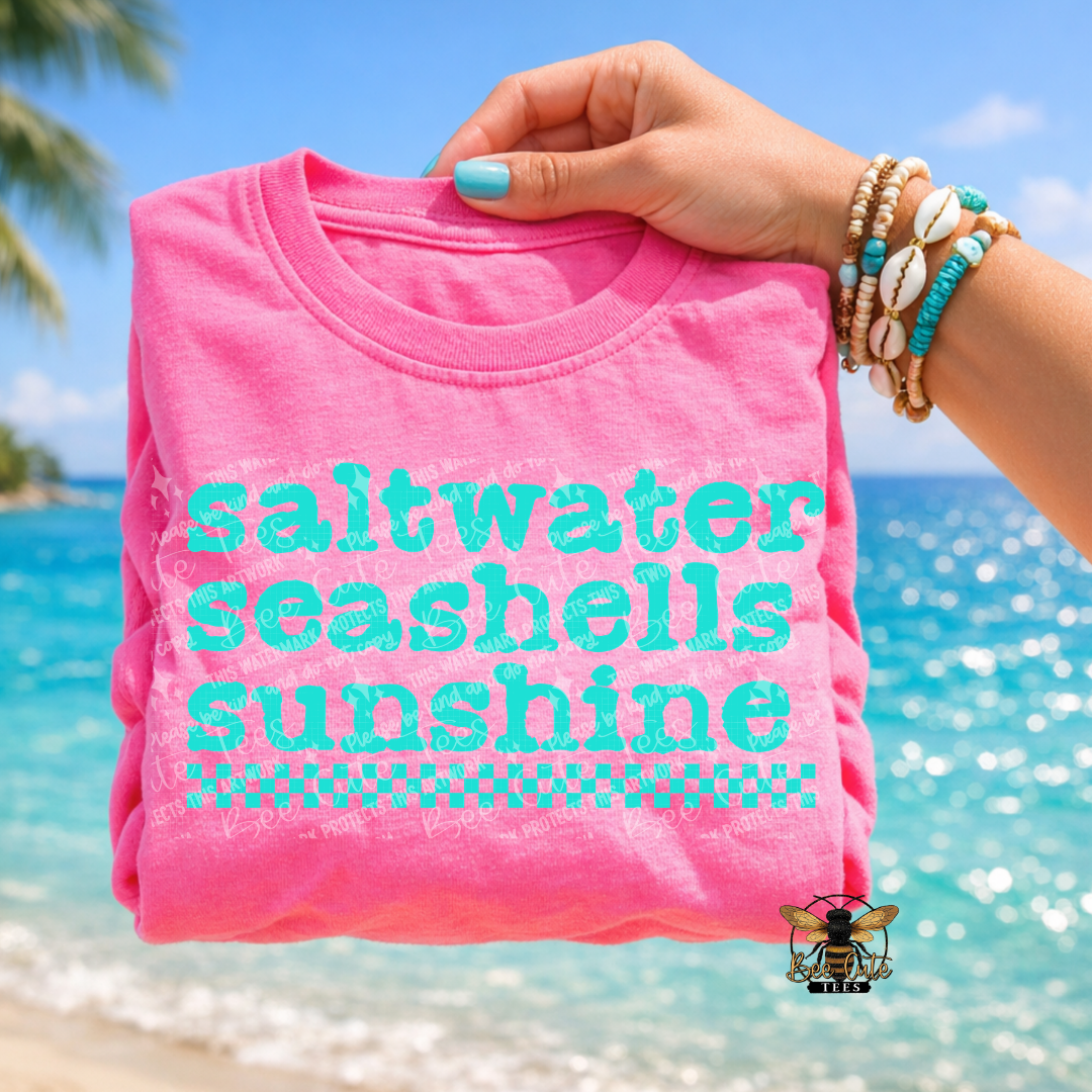 Pink t-shirt with 'saltwater seashells sunshine' text held by a hand on a beach.