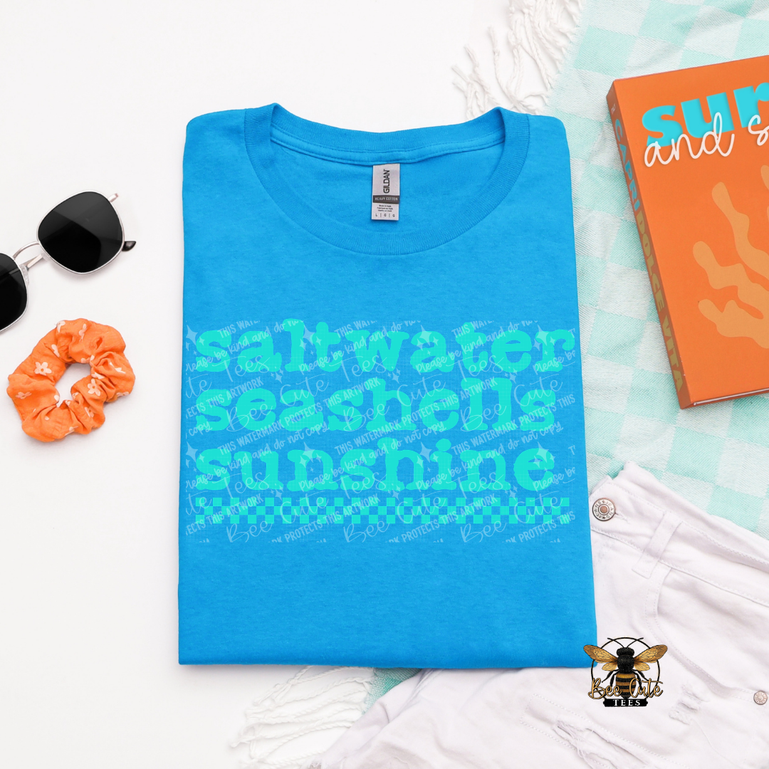 Blue t-shirt with 'saltwater seashells sunshine' text on a light blue background with accessories.