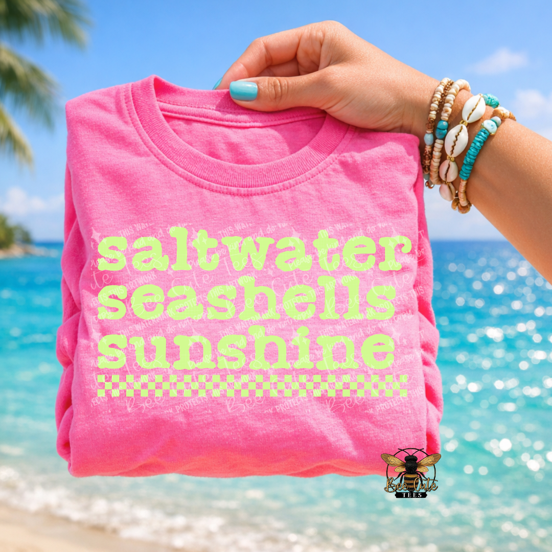 Pink t-shirt with 'saltwater seashells sunshine' text held by a hand on a beach.