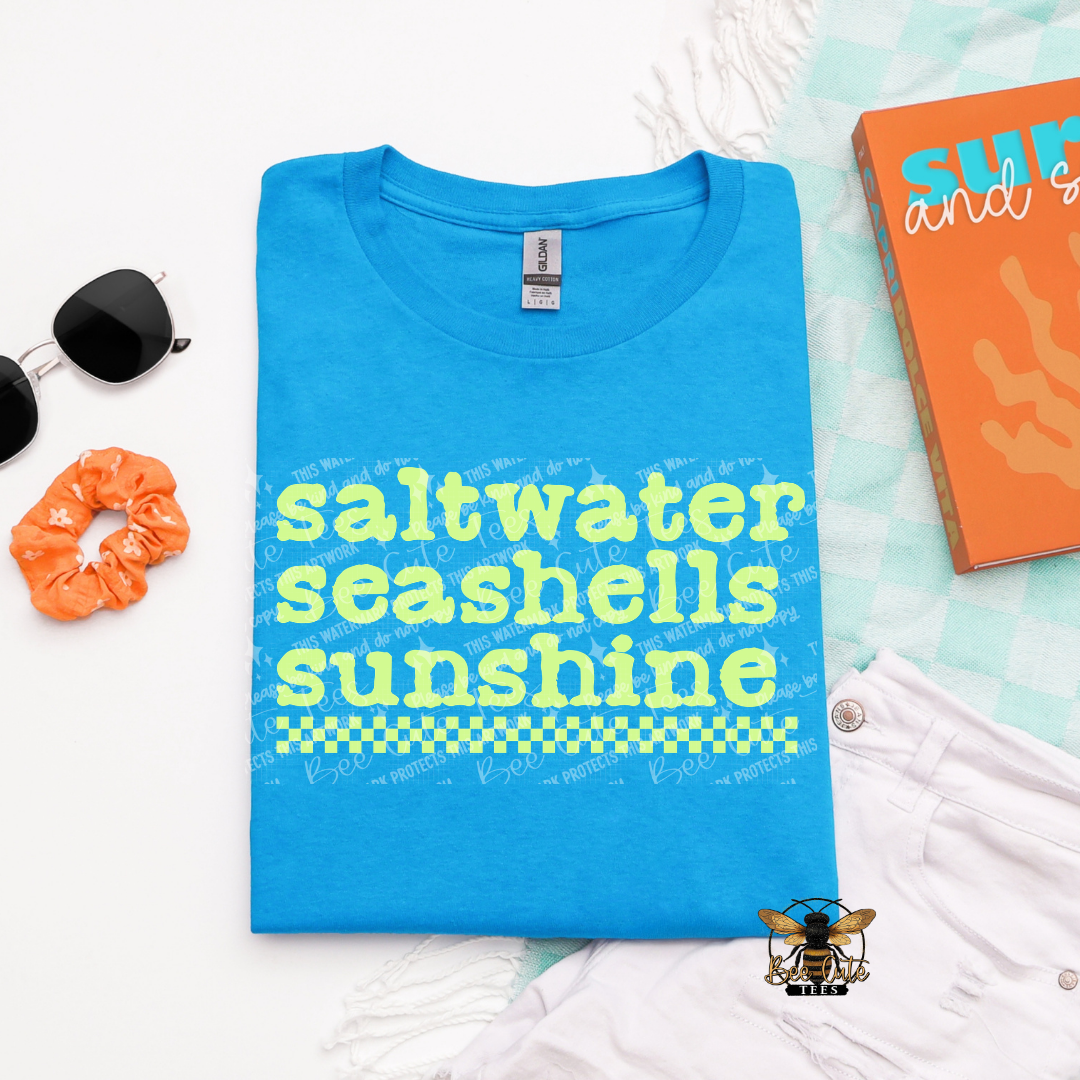 Blue t-shirt with 'saltwater seashells sunshine' text on a white surface with accessories.