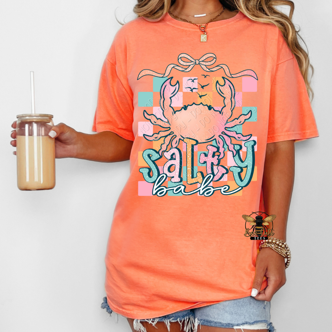Person wearing an orange t-shirt with a crab graphic and 'Salty babe' text, holding a drink.