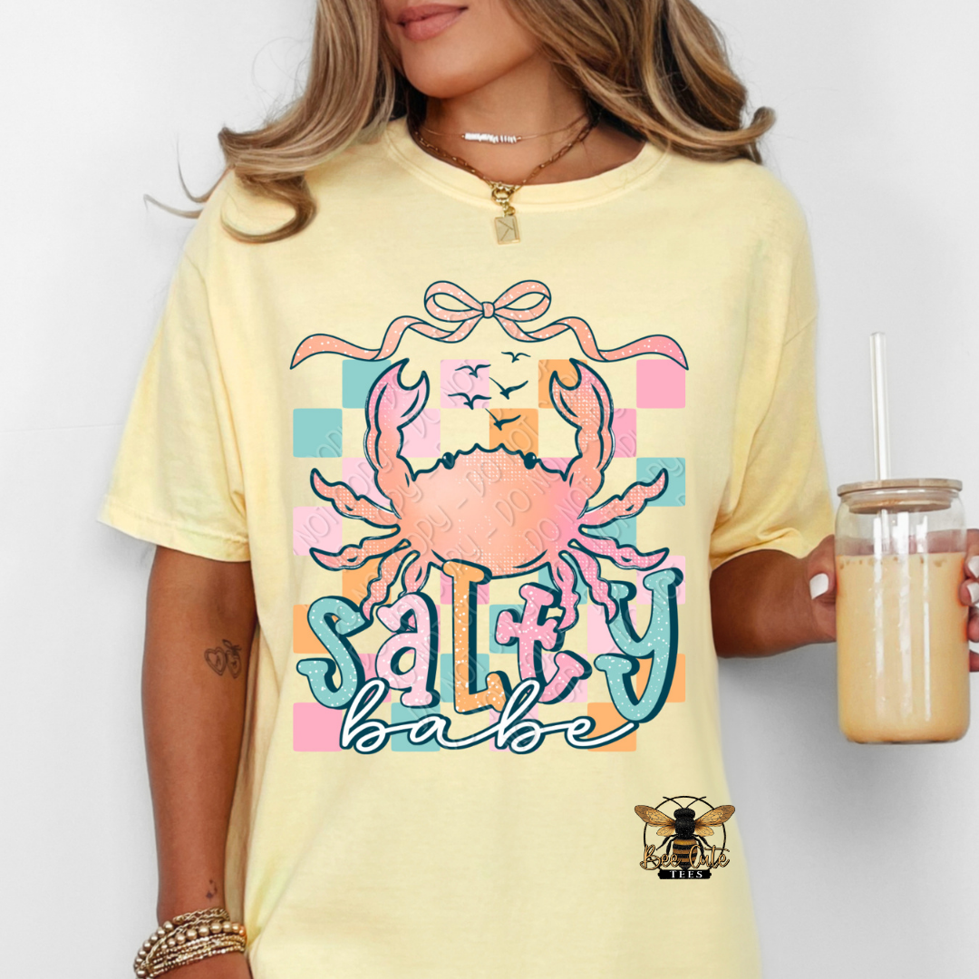 Person wearing a yellow t-shirt with a colorful crab graphic and 'Salty Babe' text, holding a drink.