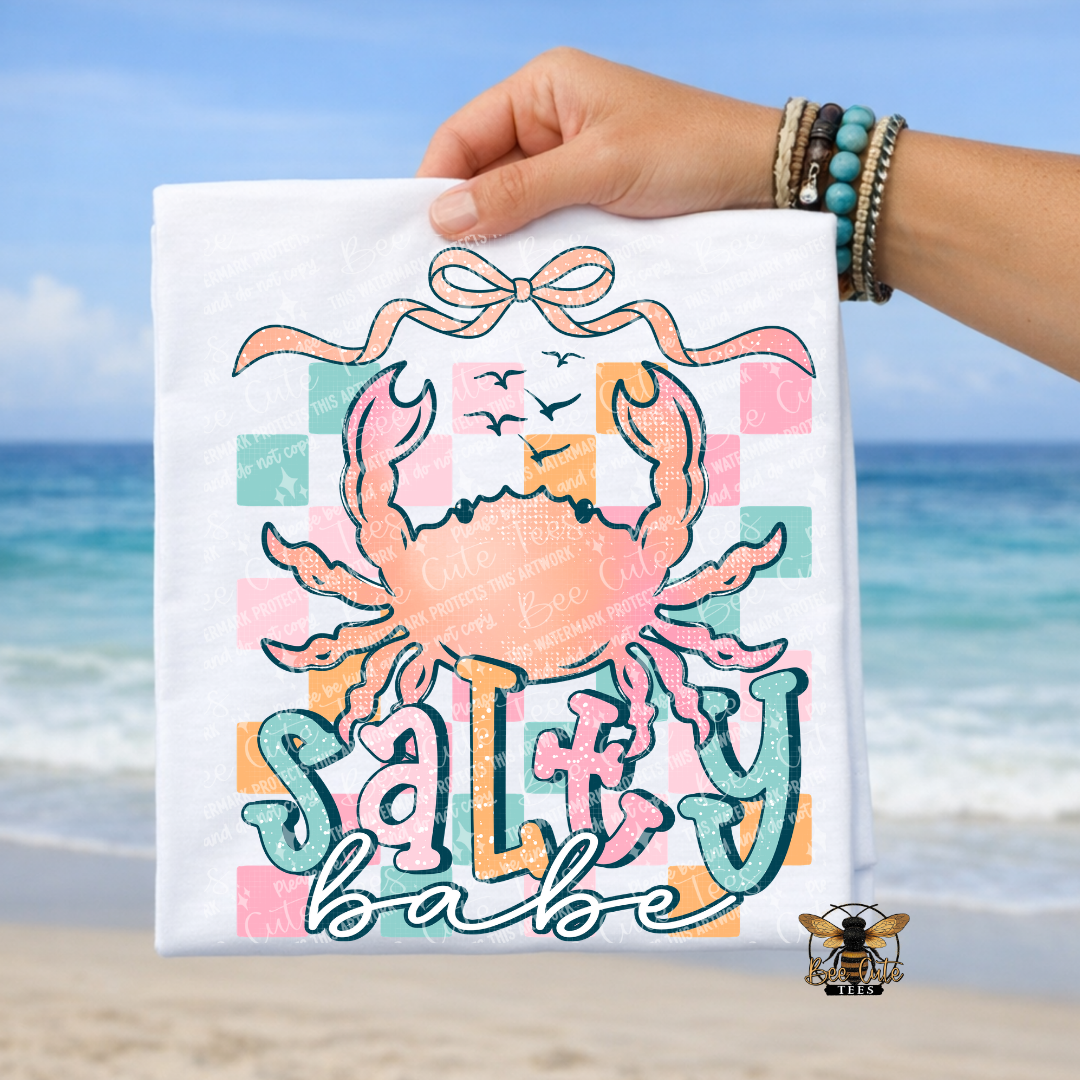 Hand holding a white t-shirt with a colorful crab design and 'Salty Babe' text against a beach background.