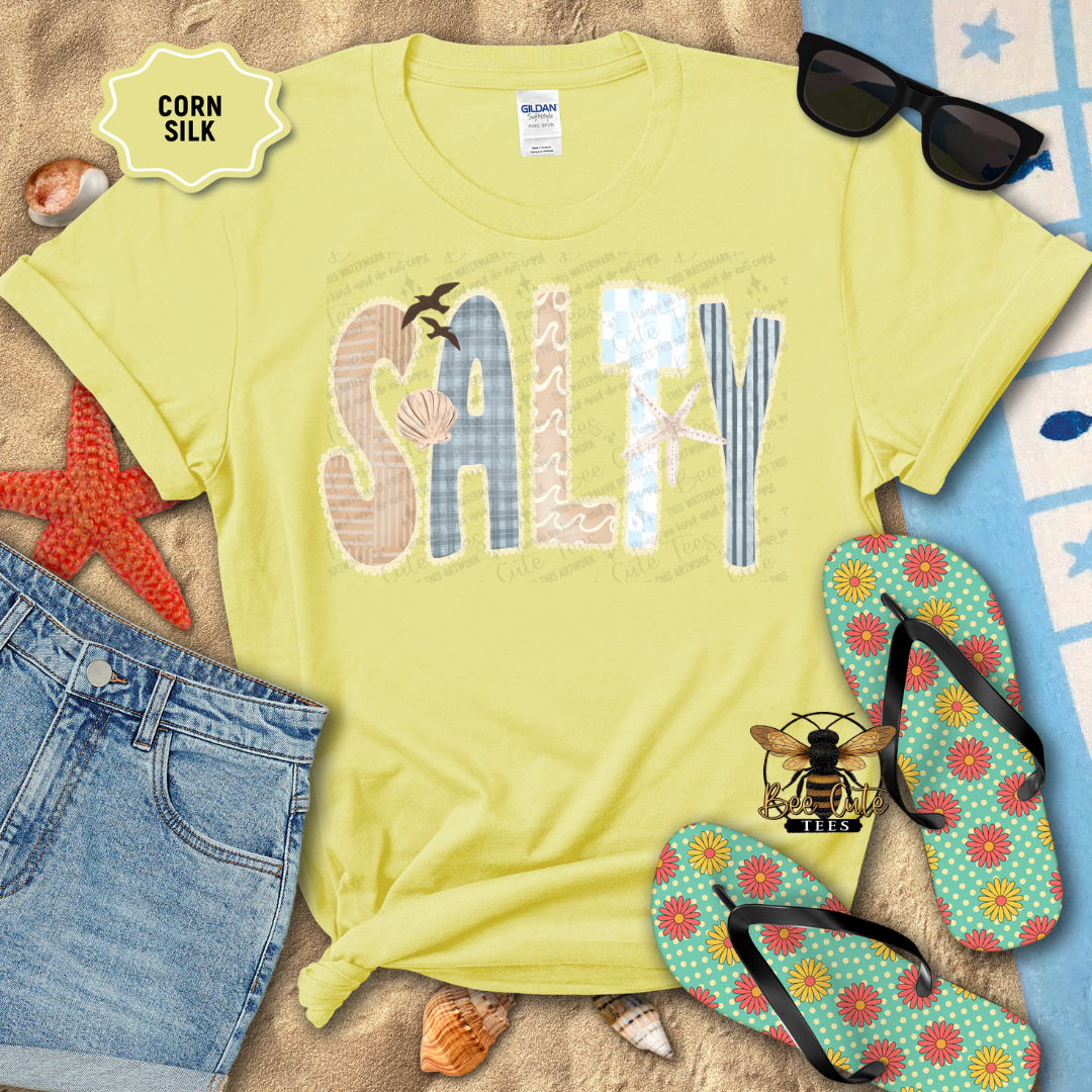 Yellow t-shirt with 'Salty' design on a sandy background with flip-flops and sunglasses.