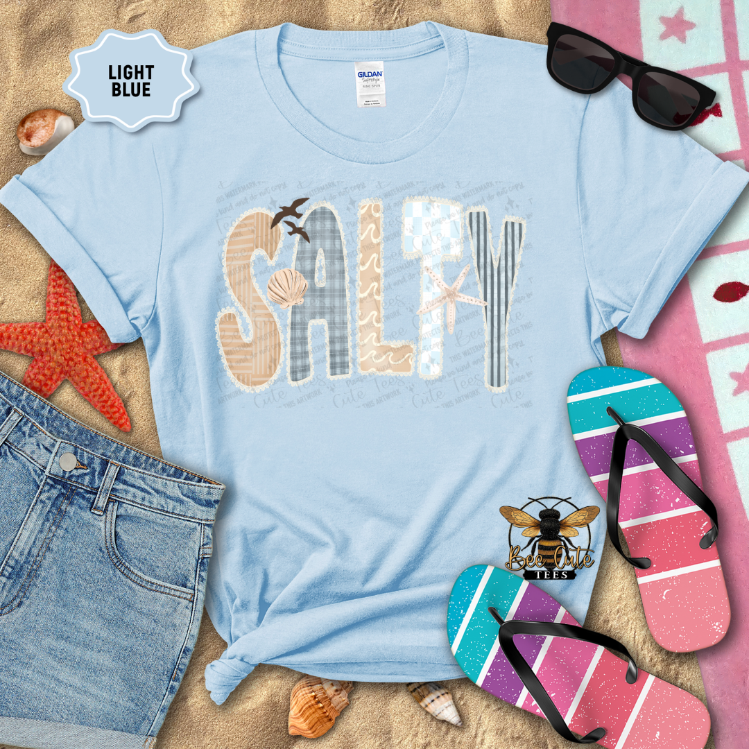 Light blue t-shirt with 'SALTY' design on a sandy background with beach accessories.
