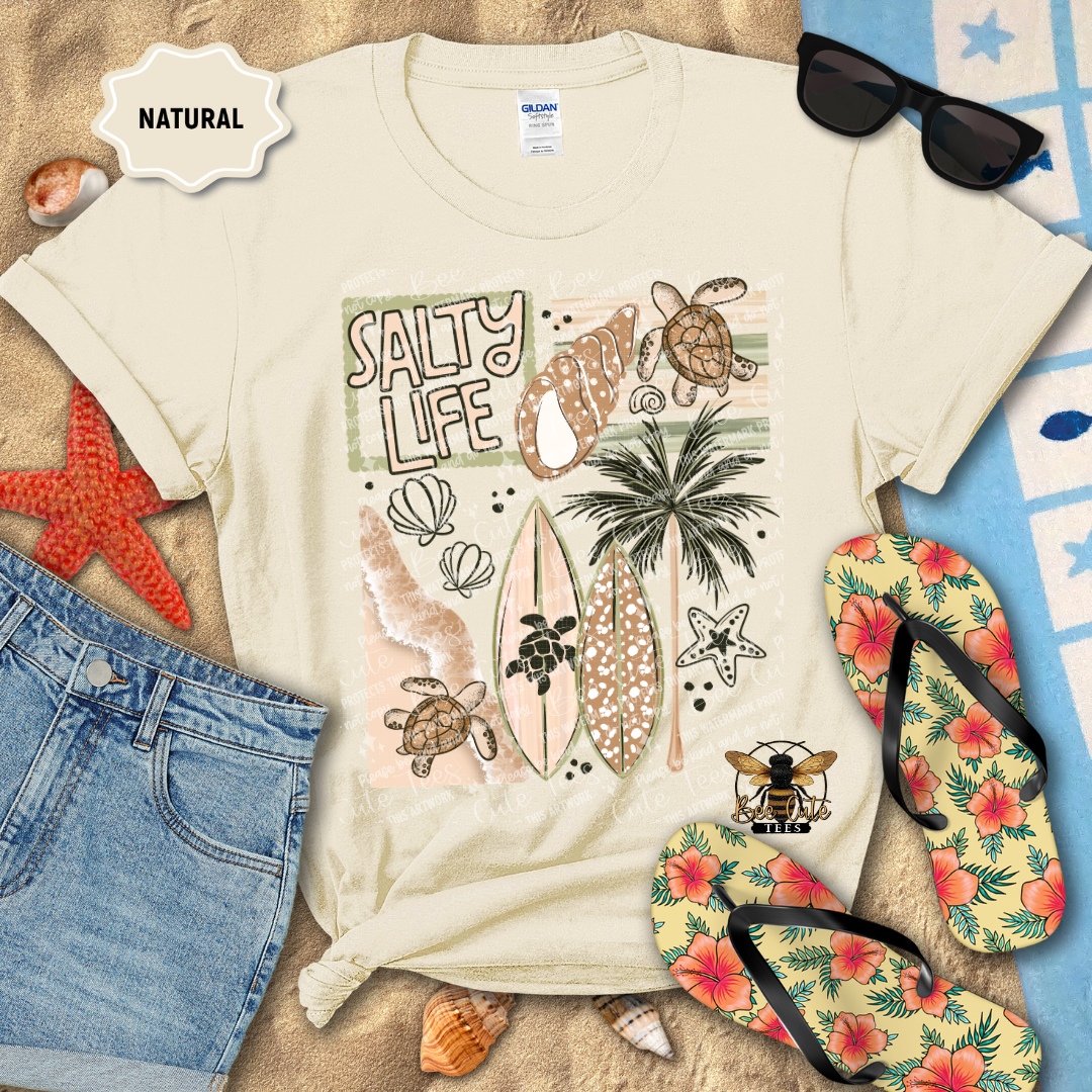 Beige t-shirt with 'Salty Life' graphic design on a sandy background with beach accessories.