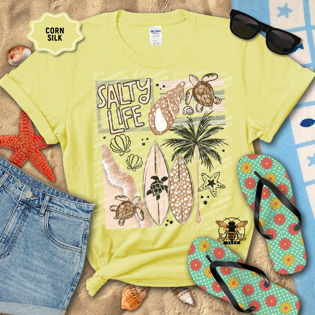 Yellow t-shirt with 'Salty Life' design, sunglasses, flip-flops, and beach accessories on a sandy background.