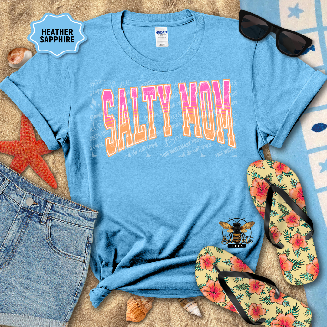 Blue t-shirt with 'Salty Mom' text, sunglasses, flip-flops, and beach items on sand.