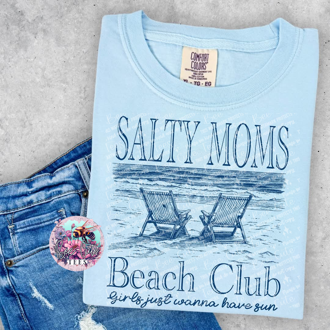 A blue t-shirt with 'SALTY MOMS Beach Club' text and an illustration of beach chairs on it, displayed flat with a pair of blue denim jeans and a decorative pin.
