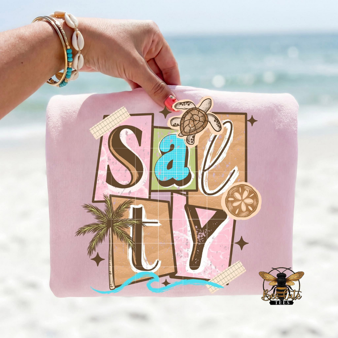 Pink t-shirt with 'Salty' design held by a hand against a beach background