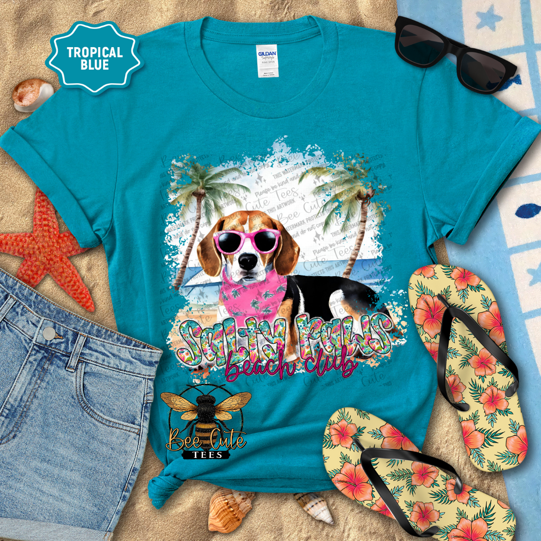 Turquoise t-shirt with a dog graphic and text, surrounded by beach accessories on sand.