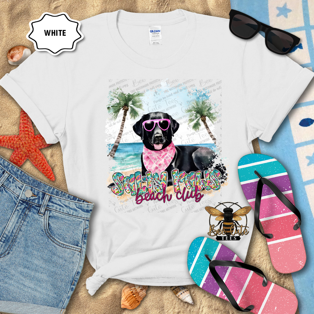 White t-shirt with a beach-themed design featuring a dog, sunglasses, and flip-flops on a sandy background.
