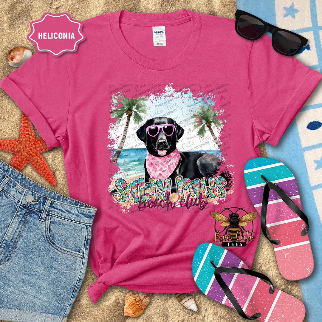 Pink t-shirt with a beach-themed design featuring a black dog, sunglasses, and flip-flops on a sandy background.