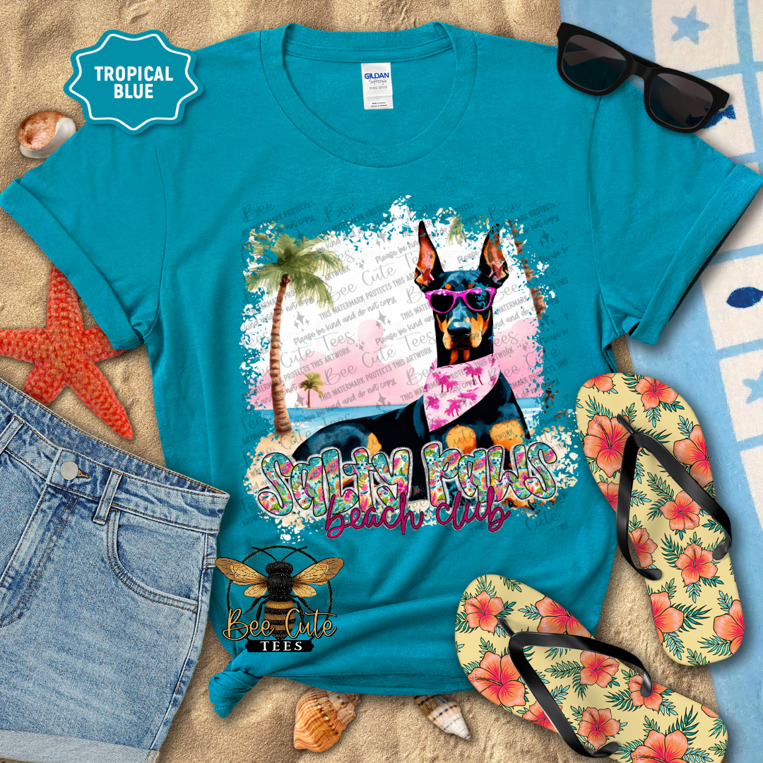 Turquoise t-shirt with a dog graphic and 'Beach Club' text, surrounded by beach accessories on sand.