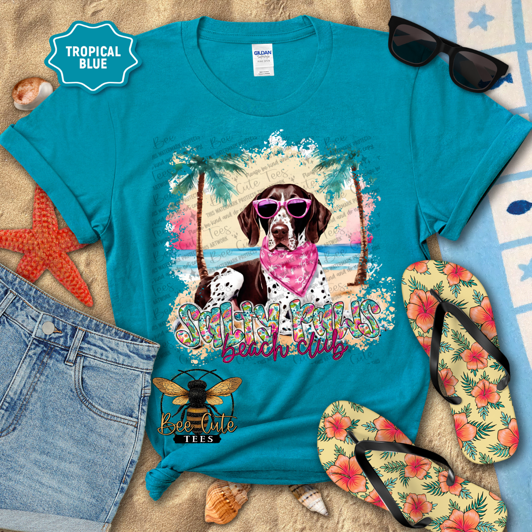 Turquoise t-shirt with a beach-themed design featuring a dog, palm trees, and text, surrounded by flip-flops and sunglasses on a sandy background.