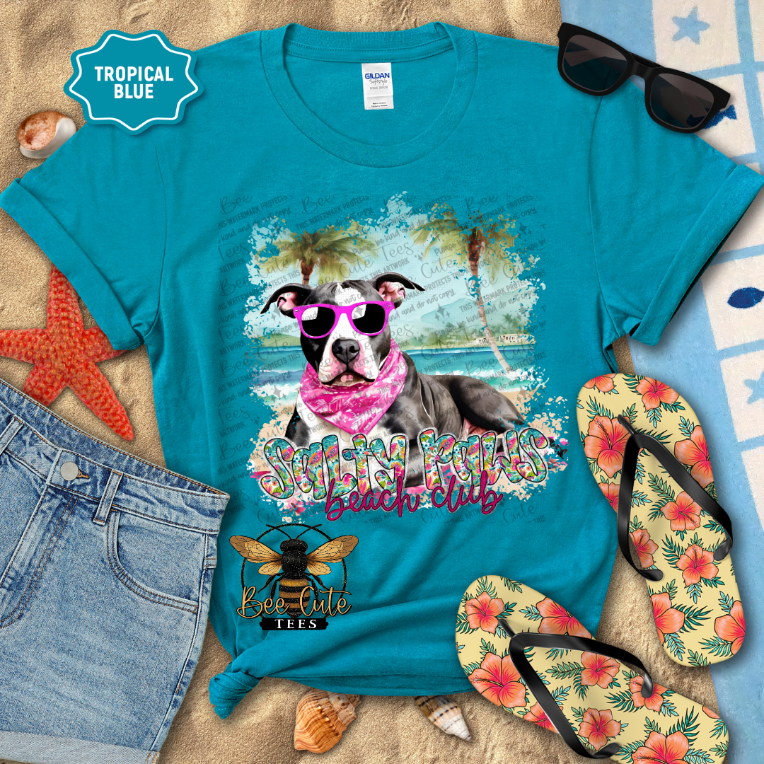 Blue t-shirt with a dog graphic and 'Beach Club' text, surrounded by beach accessories on sand.