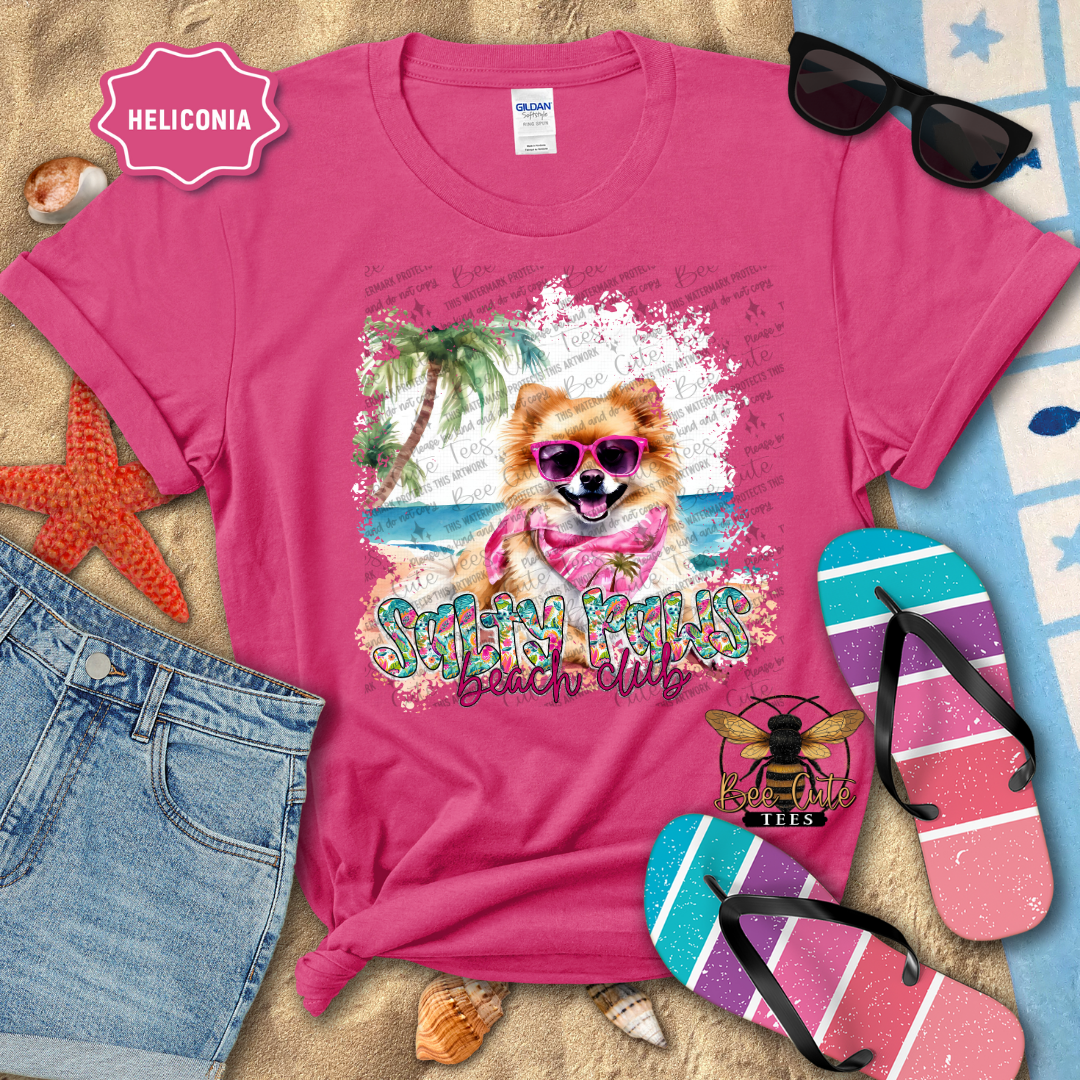 Pink t-shirt with a dog graphic and 'Swimmin' Pooches Beach Club' text, surrounded by beach accessories.