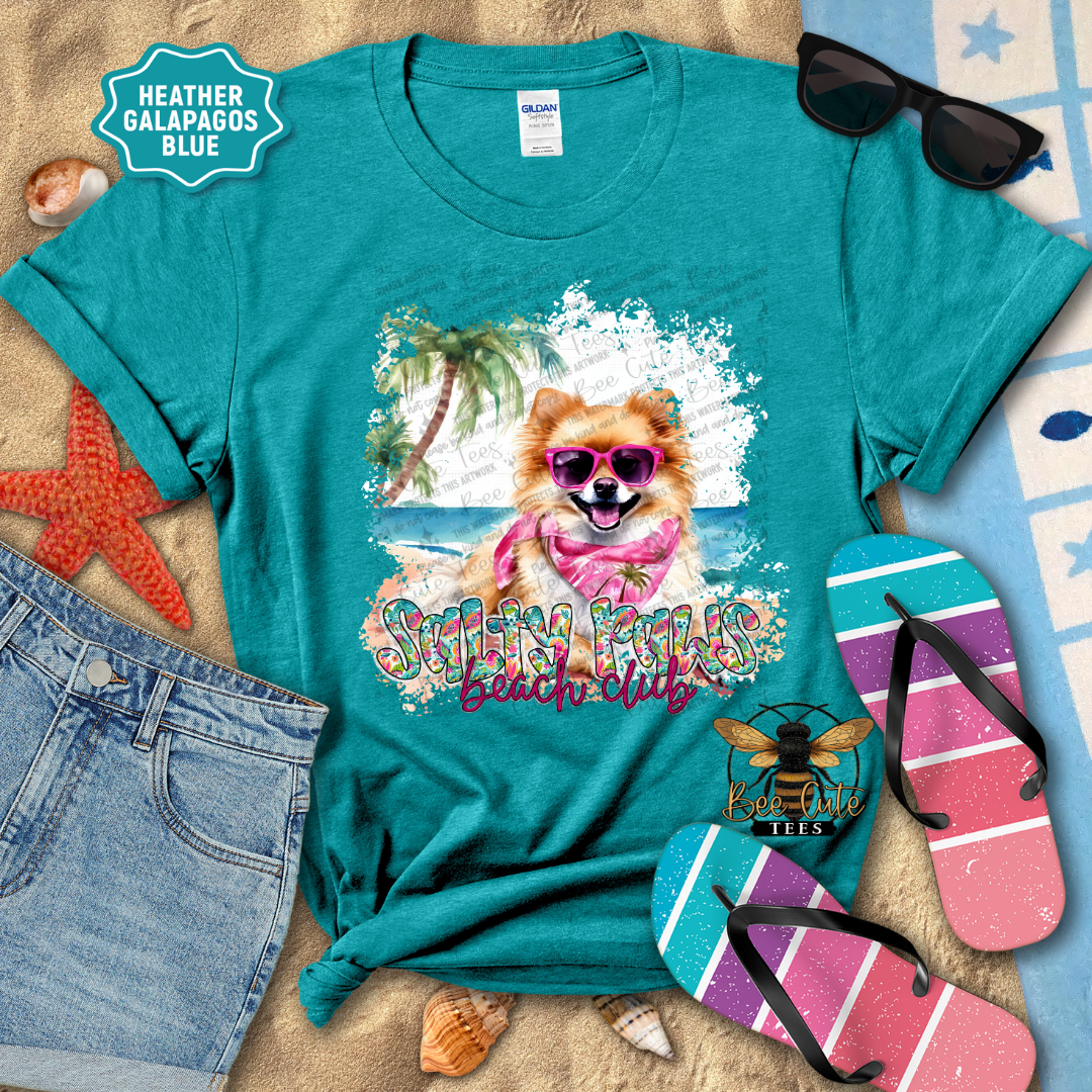 Teal t-shirt with a beach-themed design featuring a dog, sunglasses, and flip-flops on a sandy background.