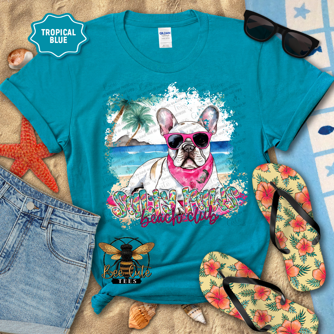Turquoise t-shirt with a beach-themed design featuring a dog, sunglasses, flip-flops, and beach accessories on a sandy background.