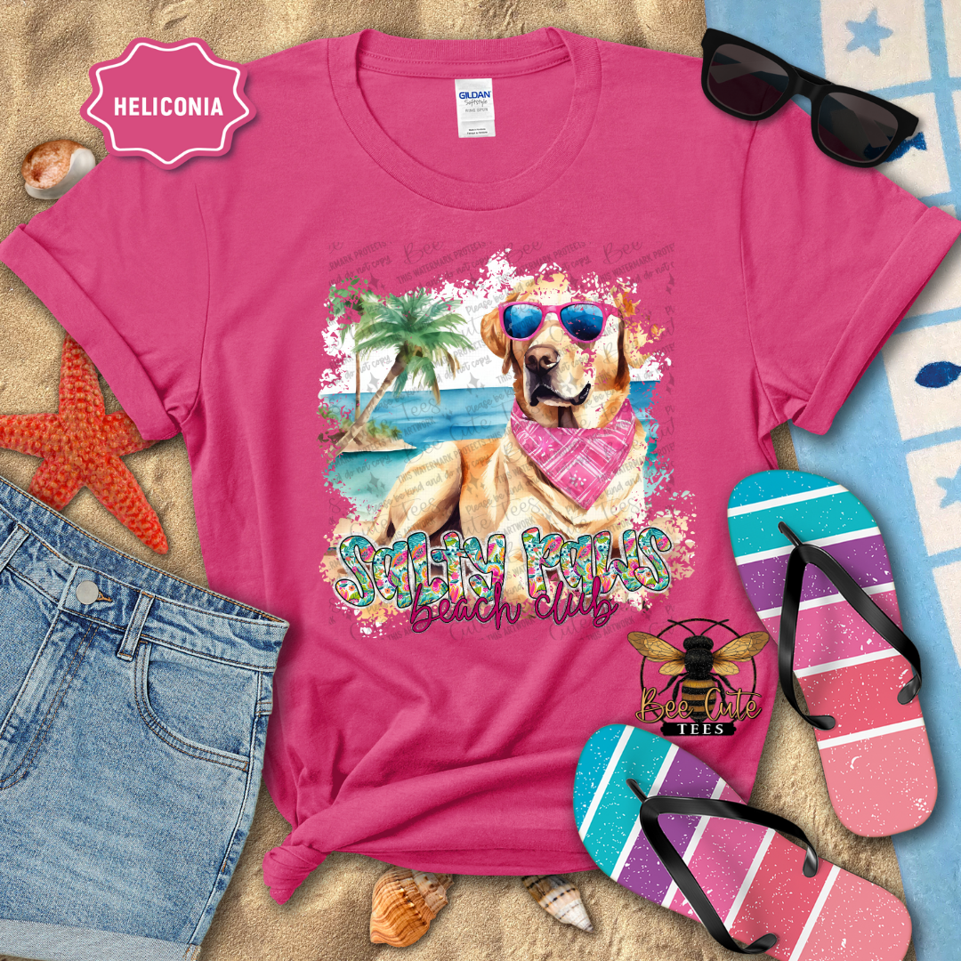 Pink t-shirt with a beach-themed design featuring a dog, sunglasses, and palm trees, surrounded by beach accessories.