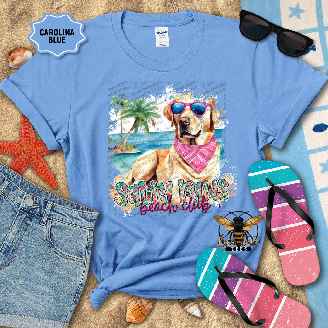 Blue t-shirt with a beach-themed design featuring a dog, sunglasses, and flip-flops on a sandy background.