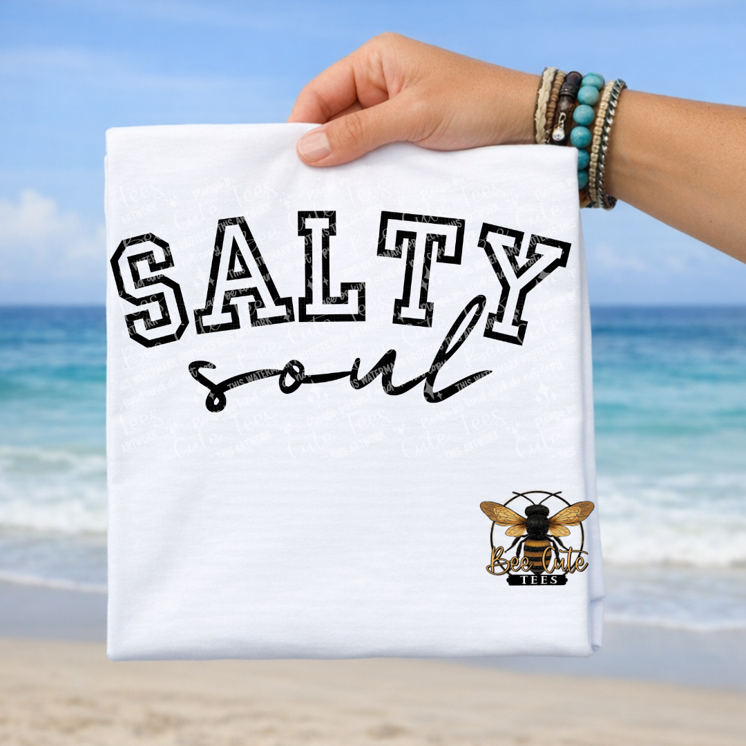 Hand holding a white t-shirt with 'Salty Soul' text and bee logo against a beach background