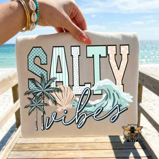 Beige t-shirt with 'Salty vibes' text and beach-themed graphics held by a hand on a wooden deck with ocean view.